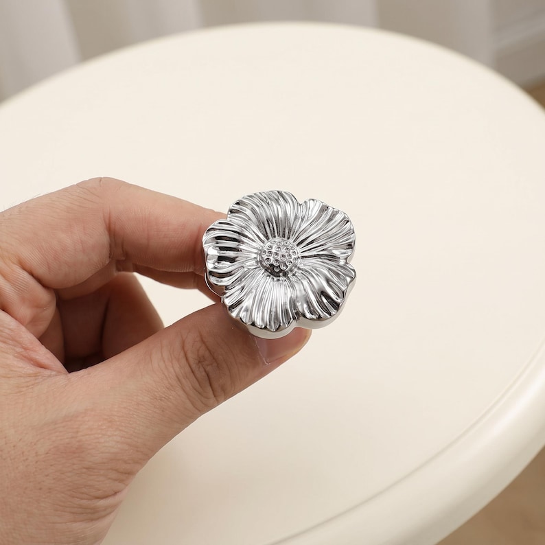 May include: A shiny silver-toned cabinet knob shaped like a flower with five distinct petals and a textured, dotted centre. The metallic surface reflects light, highlighting the radial grooves on each petal and the rounded, organic floral form.