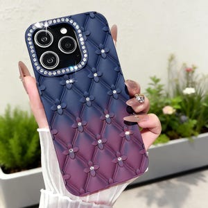 May include: A phone case with a quilted design in a gradient of blue to purple. The case features flower-shaped embellishments with embedded rhinestones and a sparkling rhinestone border around the camera lenses.