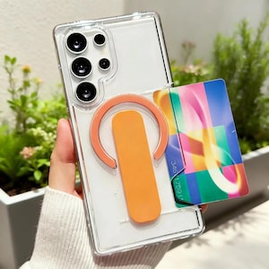 Könnte beinhalten: Transparente Handyhülle mit einem orangefarbenen Griff und einer bunten Karte. Die Handyhülle hat eine transparente Rückseite und einen transparenten Rand. Der Griff ist leuchtend orange und hat eine kreisförmige Form. Die Karte hat ein farbenfrohes Design mit der Aufschrift "346 1759".