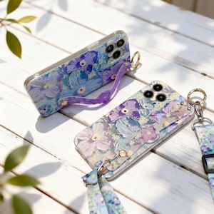 Puede incluir: Dos fundas para teléfono con diseño floral de flores moradas y azules sobre fondo claro. Cada funda tiene una correa: una morada y otra con un estampado floral a juego. Accesorios para teléfonos móviles.