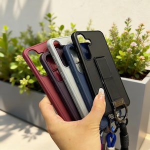 May include: A hand holding a collection of phone cases in various colors: burgundy, purple, light gray, navy blue, and black. The black case has a kickstand and a lanyard attachment. The cases are made of a smooth material.