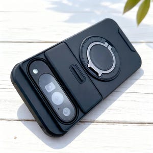 May include: A black phone case with a camera module and a silver ring holder. The case has a rectangular design with a sliding camera cover and a built-in ring for a secure grip.