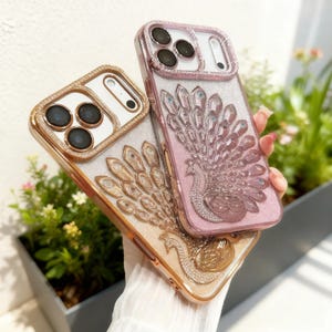 Könnte beinhalten: Zwei Handyhüllen mit gold- und rosafarbenem Glitzer-Finish. Jede Hülle hat ein detailliertes Pfauendesign mit Strassakzenten. Die Hüllen haben einen goldfarbenen Rahmen um die Kameralinsen und Kanten.