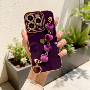 Peut inclure: Coque de téléphone violette avec un cadre et un anneau de caméra dorés. La coque est ornée d'un charm en forme de cœur doré et d'une chaîne de breloques en forme de cœur en peluche violette assortie. La coque est tenue dans une main.