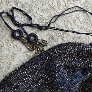 May include: Close-up of a navy blue beaded evening bag with a matching drawstring cord. The bag is intricately woven with silver beads, creating a shimmering effect. The drawstring features decorative navy blue loops and beaded tassels.