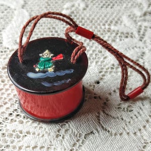 May include: A small, round, red and black container with a decorative lid featuring a painted figure in a green outfit and red scarf. The container has a brown cord handle with red accents. The item is likely a vintage trinket box or sewing accessory.