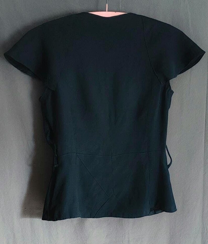 May include: Dark teal-colored top with short, flared sleeves and a V-neckline. The back of the top features a fitted waist with a subtle peplum and tie details on the sides. The top is hanging on a white hanger.