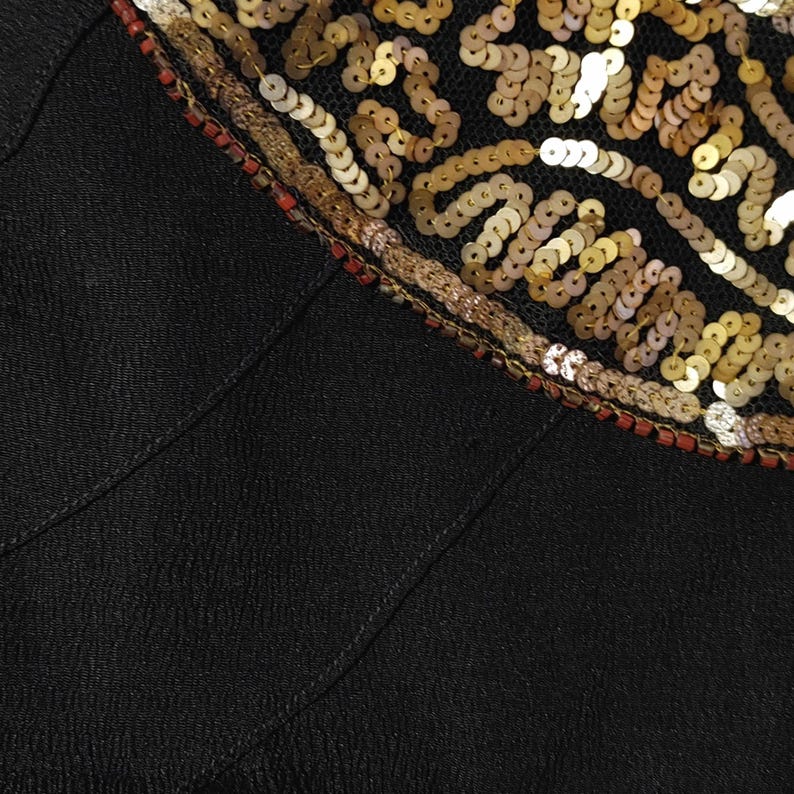 May include: Close-up of a black garment with a decorative neckline. The neckline is embellished with gold sequins, small red beads, and a gold and brown embroidered trim. The fabric has a textured appearance.