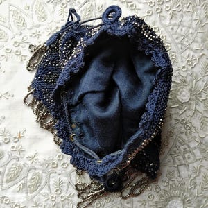 May include: A vintage navy blue beaded drawstring bag. The bag is open, revealing a dark blue interior. The exterior is heavily beaded with silver-toned beads, and the edges are trimmed with a beaded fringe. The bag has a drawstring closure.