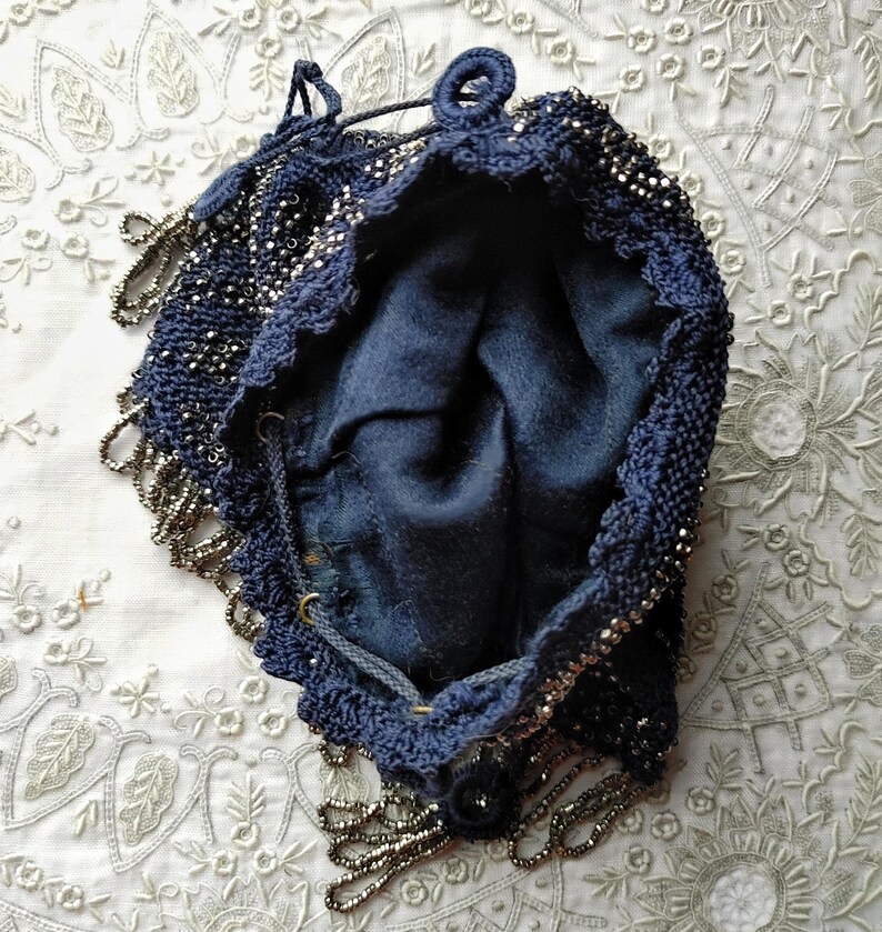 May include: A vintage navy blue beaded drawstring bag. The bag is open, revealing a dark blue interior. The exterior is heavily beaded with silver-toned beads, and the edges are trimmed with a beaded fringe. The bag has a drawstring closure.