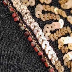 May include: Close-up of black fabric with decorative embellishments. The design includes rows of gold-colored, coin-shaped sequins and beaded strands with red and brown accents. The fabric has a textured appearance.