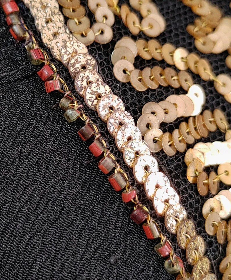 May include: Close-up of black fabric with decorative embellishments. The design includes rows of gold-colored, coin-shaped sequins and beaded strands with red and brown accents. The fabric has a textured appearance.