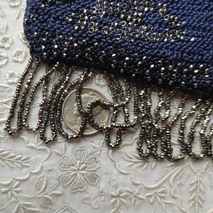 May include: Close-up of a navy blue beaded purse with a fringe of small, dark beads. The purse is heavily embellished with tiny silver beads. The fringe hangs over a quarter coin, showing the scale of the beads.