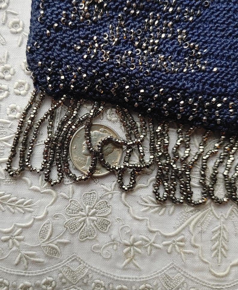 May include: Close-up of a navy blue beaded purse with a fringe of small, dark beads. The purse is heavily embellished with tiny silver beads. The fringe hangs over a quarter coin, showing the scale of the beads.