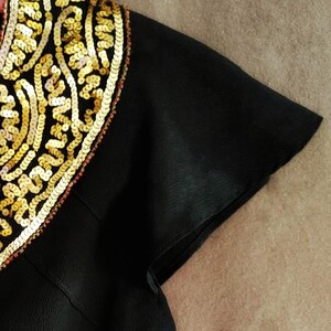 May include: Close-up of a black garment with a short sleeve. The shoulder area is adorned with intricate gold sequins in a decorative pattern. The sequins are outlined with a thin red trim.