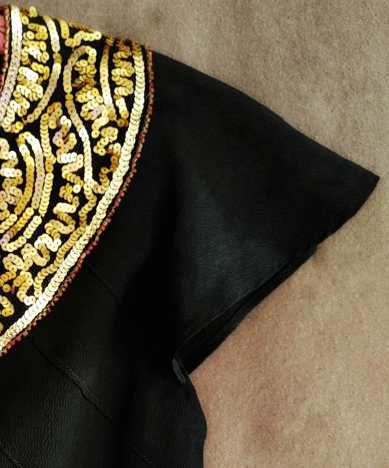 May include: Close-up of a black garment with a short sleeve. The shoulder area is adorned with intricate gold sequins in a decorative pattern. The sequins are outlined with a thin red trim.