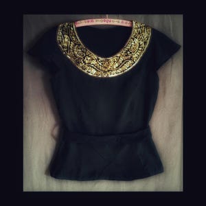 May include: Black short-sleeved top with a gold sequined collar and a matching belt. The collar features an intricate design. The top is hanging on a pink hanger. The waistline has a diamond pattern.