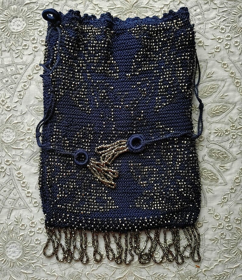 May include: A navy blue beaded handbag with a drawstring closure. The bag features a decorative beaded design and a beaded fringe along the bottom. The bag has a strap with two circular accents and a beaded tassel.