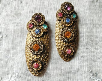 May include: A pair of oval-shaped earrings with a hammered gold-tone metal finish. Each earring features multiple floral-shaped settings containing small, multi-colored rhinestones in shades of pink, blue, orange, and purple. The text IN THE RUINS is visible.