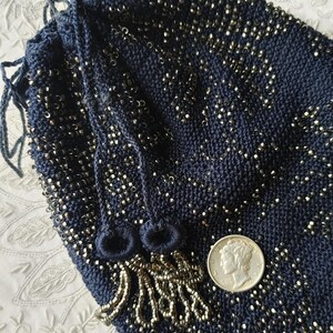 May include: A navy blue drawstring bag, heavily embellished with silver-colored beads. The bag's knitted texture is visible, and it features a beaded fringe and drawstring closure. A silver coin is placed next to the bag for size comparison.