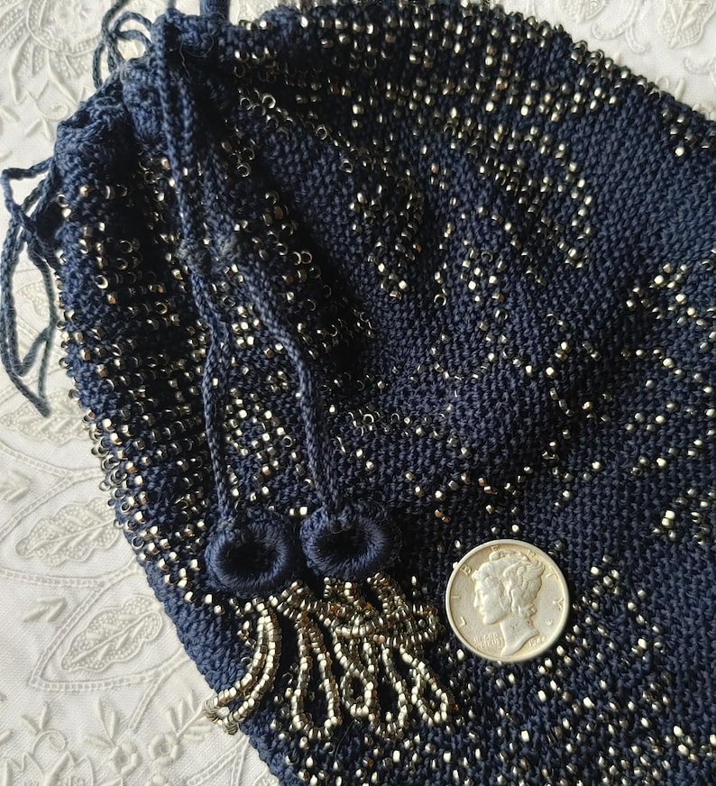 May include: A navy blue drawstring bag, heavily embellished with silver-colored beads. The bag's knitted texture is visible, and it features a beaded fringe and drawstring closure. A silver coin is placed next to the bag for size comparison.