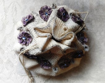 May include: A cream-colored crocheted drawstring pouch featuring a ruffled top edge and dark purple fabric flower embellishments with small blue center beads. The text "IN THE RUINS" is printed in a simple font at the bottom of the image.
