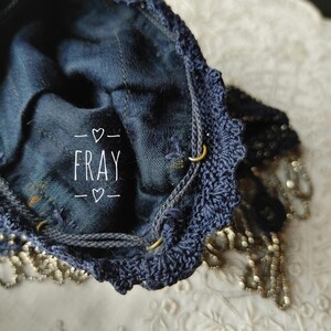 May include: A dark blue drawstring bag with a crocheted edge and metal ring details. The word "FRAY" is printed in white. The bag is made of a denim-like material and has a decorative beaded fringe.