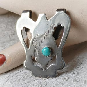 May include: A silver-toned money clip with an engraved design and a turquoise stone. The clip has a decorative shape with the name "Lumia" engraved on it. The turquoise stone is set in the center of the clip.