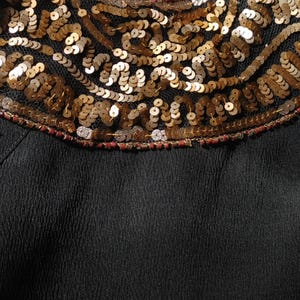 May include: Close-up of a black garment with a decorative neckline. The neckline features intricate gold sequins and beading, creating a textured, ornate design. The sequins are arranged in a swirling pattern against a sheer black fabric.