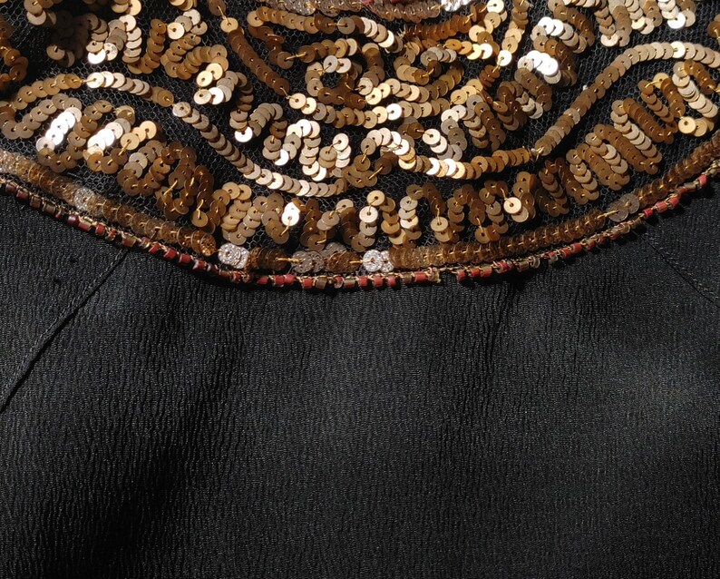 May include: Close-up of a black garment with a decorative neckline. The neckline features intricate gold sequins and beading, creating a textured, ornate design. The sequins are arranged in a swirling pattern against a sheer black fabric.