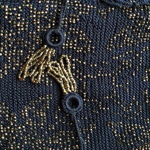 May include: Close-up of a navy blue knit fabric with gold metallic threads. The fabric features a beaded design in gold, with two navy blue buttons and a beaded loop closure. The texture is detailed and the overall look is elegant.