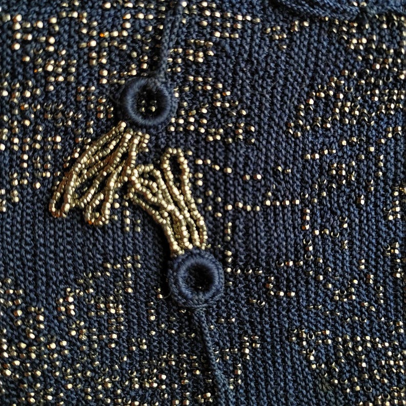 May include: Close-up of a navy blue knit fabric with gold metallic threads. The fabric features a beaded design in gold, with two navy blue buttons and a beaded loop closure. The texture is detailed and the overall look is elegant.