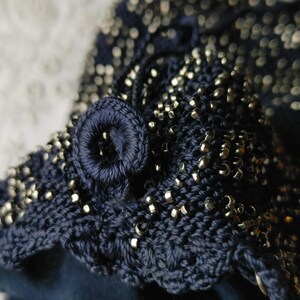 May include: Close-up of a navy blue crocheted item with silver-colored beads. The item features a scalloped edge and a circular, embroidered detail. The beads are evenly distributed across the surface, adding texture and shine.