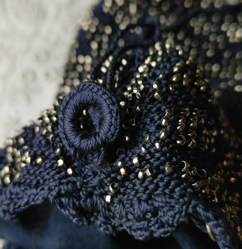 May include: Close-up of a navy blue crocheted item with silver-colored beads. The item features a scalloped edge and a circular, embroidered detail. The beads are evenly distributed across the surface, adding texture and shine.