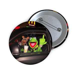Op de afbeelding: Een ronde button met Kermit de Kikker in een auto, die de middelvinger opsteekt. Miss Piggy zit op de passagiersstoel. Een McDonald's-bord is zichtbaar op de achtergrond. De button is zwart met een zilveren speld.