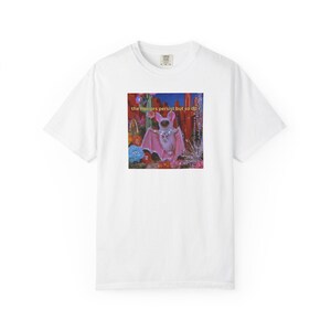 Peut inclure: T-shirt blanc avec un motif d'une chauve-souris rose avec un collier et des ailes ornés de bijoux, sur un fond de paysage floral et désertique coloré. Le texte "the horrors persist but so do i" est au-dessus de la chauve-souris.