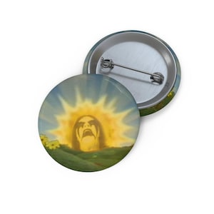 May include: Two round metal pinback buttons. One button features a sun with a face rising over a green hill. The sun has a face with dark hair and a screaming mouth. The other button is silver.