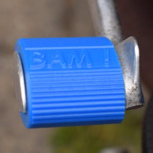 Könnte beinhalten: Ein leuchtend blaues, zylindrisches Pedal mit gerippter Textur und dem eingeprägten Wort BAM an der Seite. Das Pedal ist an einem silberfarbenen Metallarm befestigt und verfügt über eine glatte, abgerundete Endkappe sowie eine kleine Metalllasche.