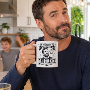 May include: A white ceramic mug with the text "I'm Not Ignoring You, I'm Processing in Dad Silence" and a black and white illustration of a man. The mug is held by a person wearing a navy blue long-sleeve shirt.