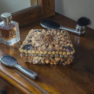 May include: A decorative box, encrusted with seashells, rests on a wooden surface, alongside a perfume bottle and two hairbrushes. The box is covered in a variety of shells in shades of tan, brown, and cream. The box is approximately 15 cm square.