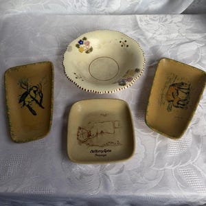 Puede incluir: Un conjunto de cuatro piezas de cerámica: un cuenco y tres bandejas pequeñas. El cuenco es de color crema con detalles florales. Las bandejas son de color beige con diseños pintados, incluyendo un pájaro, un oso y un edificio con el texto "Old Harry Rocks, Swanage."