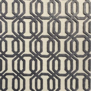 May include: A rectangular rug with a repeating geometric pattern. The design features interlocking dark grey shapes against a light beige background. The rug's texture appears soft and plush, suitable for a living room or bedroom.