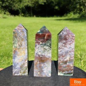 May include: Three polished gemstone obelisks with pointed tops. The stones display a mix of colors, including red, green, white, and brown. The obelisks are standing on a black surface with a green grassy background.