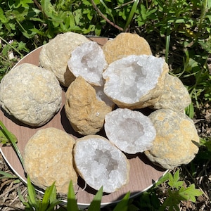 Peut inclure: Une collection de roches géodes rondes de couleur beige avec des extérieurs rugueux et texturés. Plusieurs pierres sont fendues pour révéler des centres creux remplis de grappes de petits cristaux de quartz blancs, clairs et étincelants.