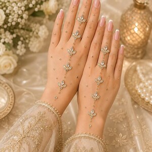 May include: A gold-toned hand jewelry piece featuring a series of marquise-cut clear crystals arranged in a vertical line, connected by delicate chains and small gold beads, extending from the fingers down to the wrist over a sheer lace sleeve.
