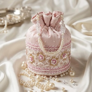 Puede incluir: Un bolso redondo rosa claro con cordón, con intrincados bordados florales y abalorios. El bolso está adornado con perlas y un asa de cuentas. El fondo es una tela de satén blanco, con perlas dispersas.
