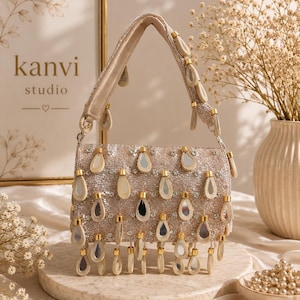 May include: A beige handbag featuring a dense surface of small beads and numerous dangling teardrop-shaped mirror accents. The bag includes a matching shoulder strap adorned with similar mirror charms and a secure top closure.