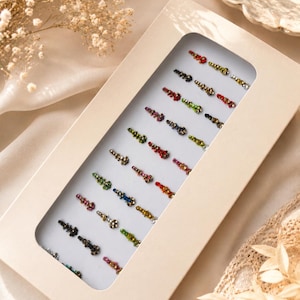 Puede incluir: Una tarjeta de presentación rectangular de cartón blanco que contiene una colección de pequeños adhesivos bindi multicolores. Las pegatinas están dispuestas en filas verticales y presentan varias formas, incluyendo rojo, azul, verde, oro y negro.