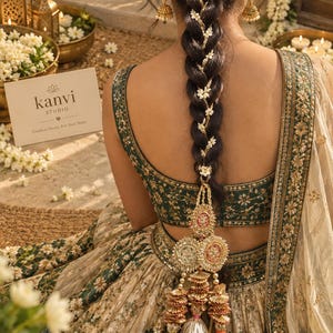 May include: A dark-haired person wears a green and cream embroidered lehenga with a long, thick braid adorned with small white flowers. A decorative gold-toned hair accessory with circular pendants and metallic tassels hangs from the base of the braid.