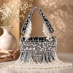 May include: A black handbag featuring an intricate exterior adorned with white pearls and clear crystals. The bag includes a top handle embellished with pearls and a long, cascading fringe of white pearls hanging from the base and handle.
